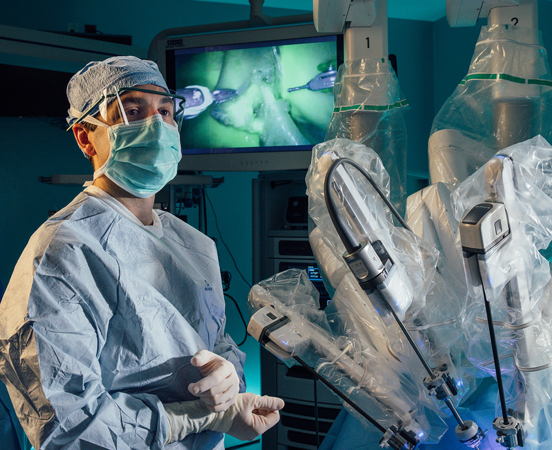 General surgeon Dr. Giannone stands in full PPE beside the robotic arms of a surgical robot that he'll control during surgery.